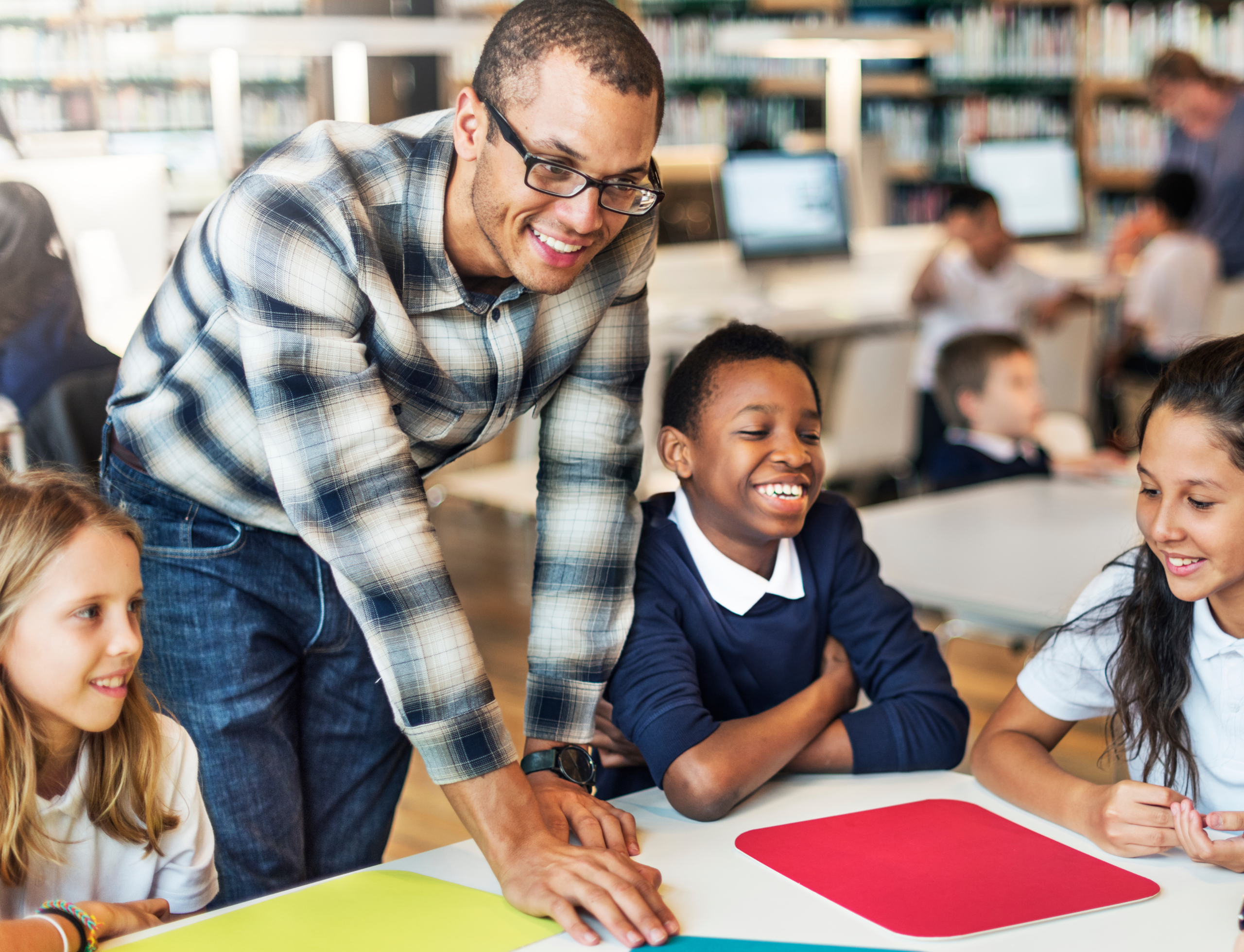 Smiling teacher leaning over a table to help three primary school pupils in a school library